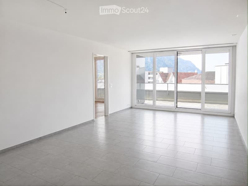Empty room with white walls, grey tiled floor, large sliding glass doors leading to a balcony, and a view of mountains and buildings.