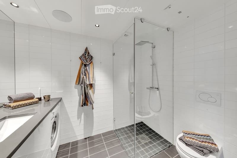 Modern bathroom with white tiled walls, a glass-enclosed shower, a sink with a mirror, a toilet, and a washing machine. Towels and a bathrobe are neatly arranged on the counter and toilet seat.