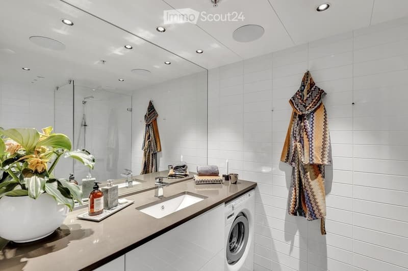 Modern bathroom with white tiles, double sink, large mirror, towel rack, and a washing machine. Features a decorative plant and toiletries on the counter.