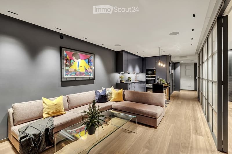 Modern living room with a sectional sofa, glass coffee table, potted plant, and artwork. Adjacent kitchen area features dark cabinets, a sink, microwave, and pendant lights.