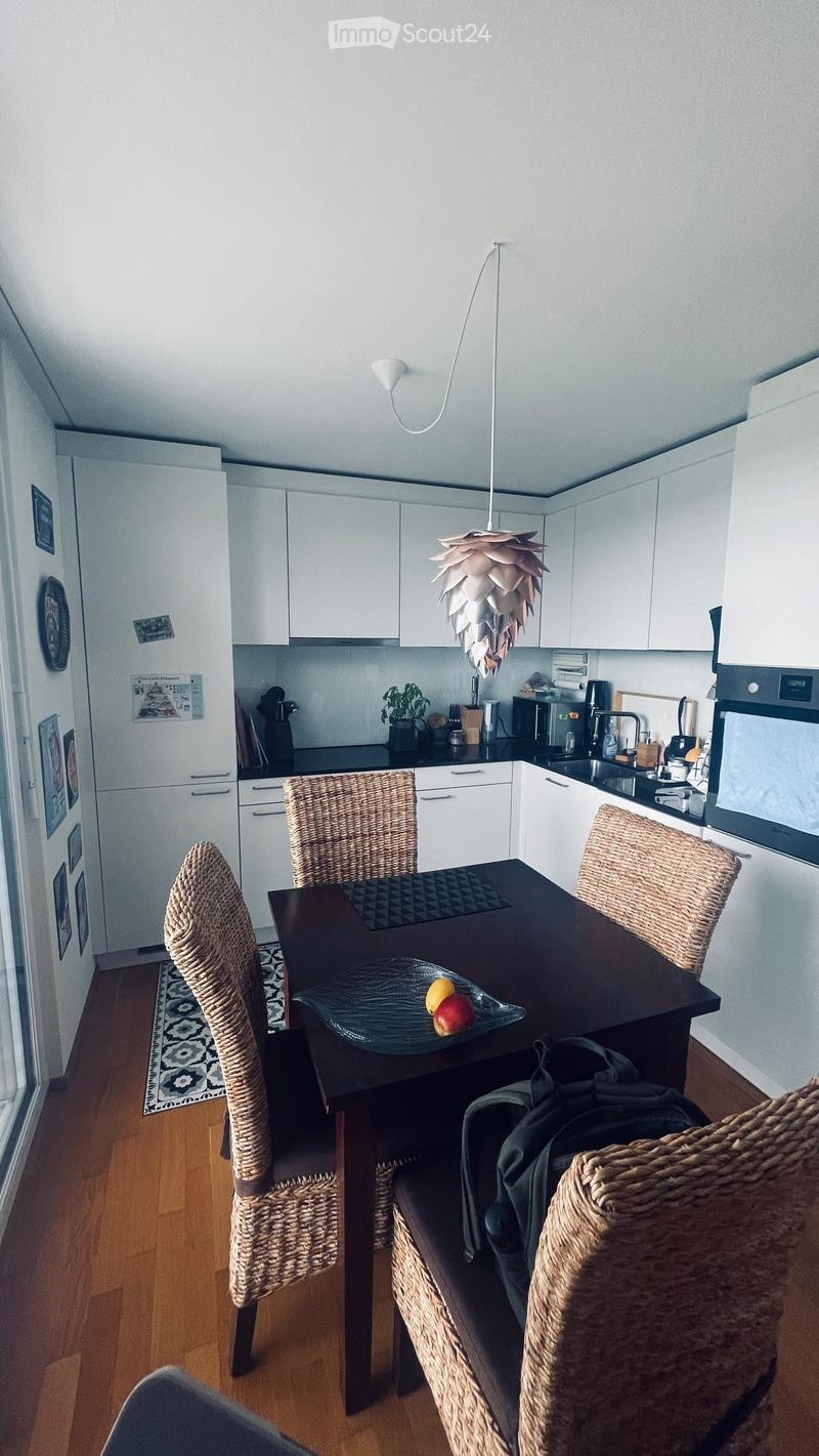 Modern kitchen with white cabinets, black countertops, and a dark wooden dining table surrounded by wicker chairs. A fruit bowl with apples and oranges sits on the table, and a backpack is placed on one of the chairs. The kitchen features a pendant light,