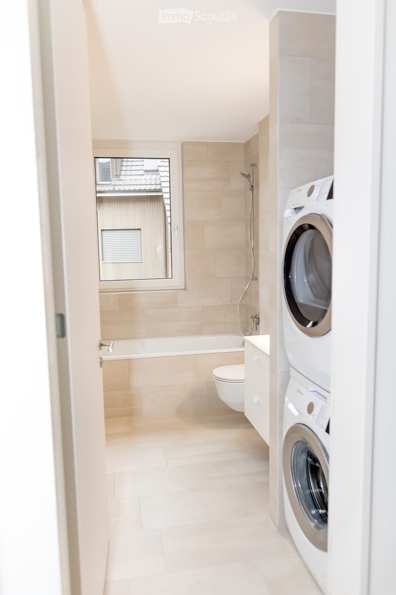 Modern bathroom with beige tiles, a bathtub, toilet, washing machine, and a large mirror above the window.