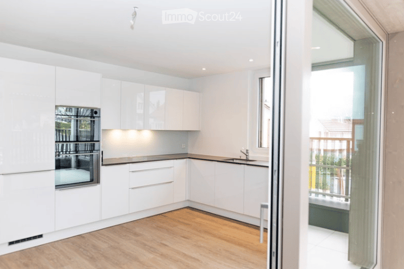 Modern, minimalist kitchen with white cabinetry, a built-in oven, and a sleek sink. The space features light wood flooring and an open sliding door leading to a balcony.