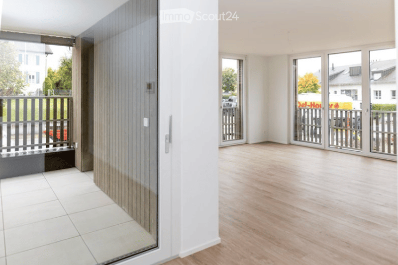 Bright, empty room with wooden flooring and large glass doors leading to a balcony area. The space is clean and minimalistic, with no furniture or decorations.