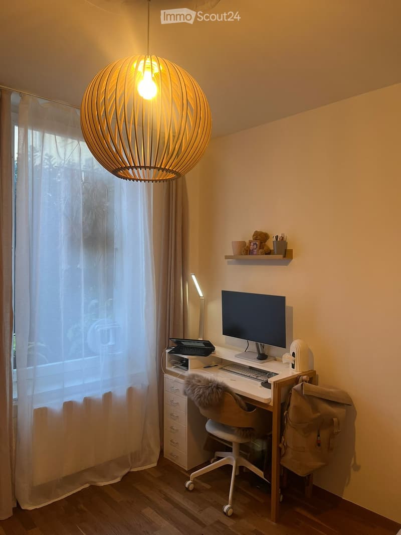 A modern, well-lit workspace with a wooden desk, ergonomic chair, laptop, monitor, keyboard, mouse, and a decorative pendant light. The room features large windows with sheer curtains and a small shelf with decorative items.