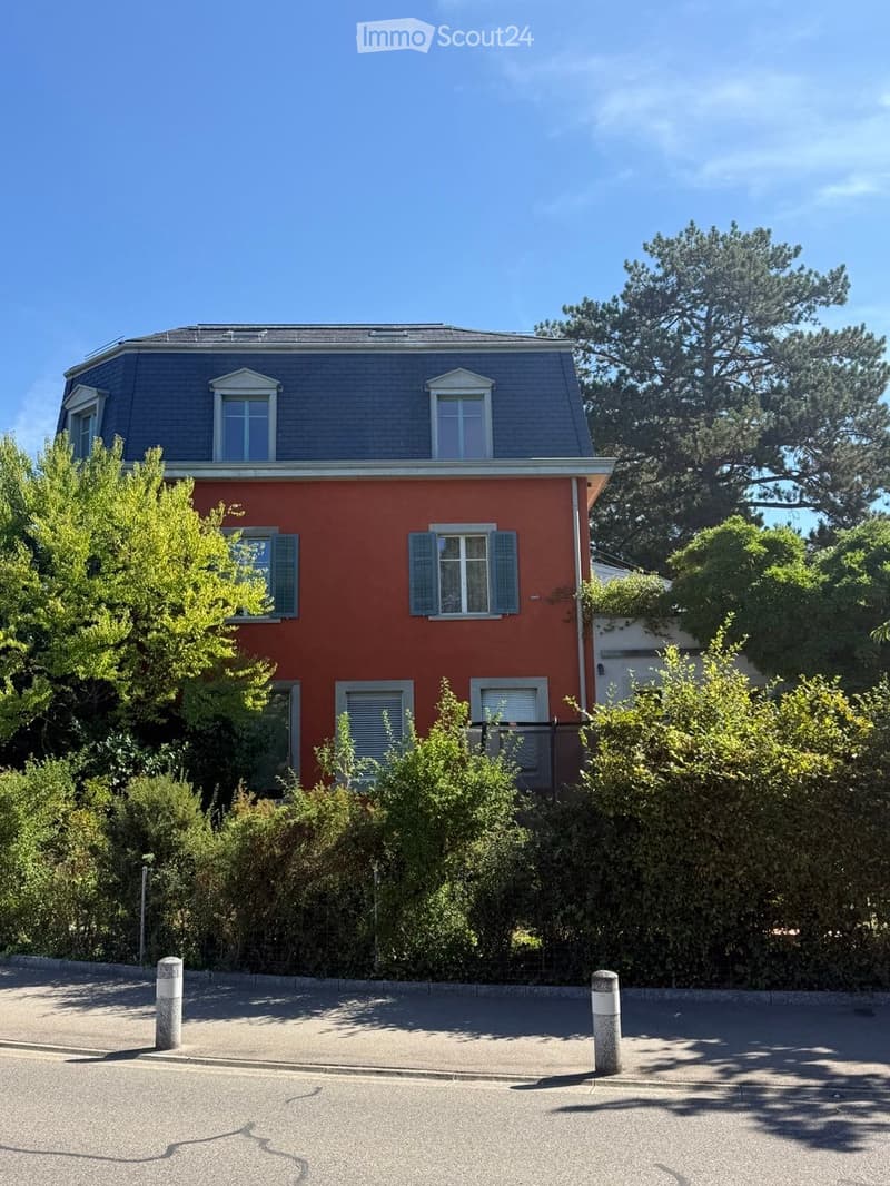 Two-story red house with grey roof, multiple windows, and surrounding greenery.