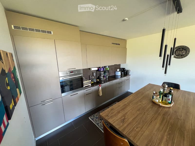 Modern kitchen with light grey cabinets, stainless steel oven, sink, and coffee maker. Wooden dining table with chairs and various bottles on it.