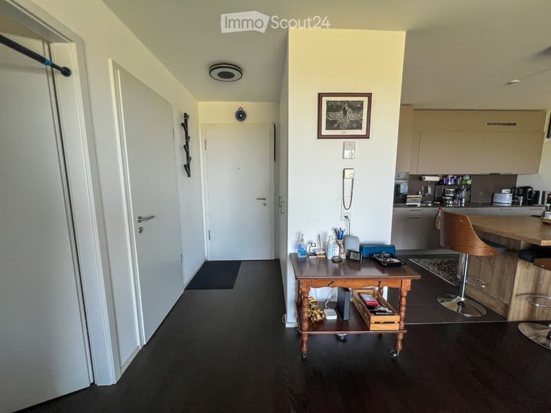 A modern, well-lit hallway leading to a kitchen area. The hallway features a dark wooden floor, white walls, and a small wooden table with various items on it. The kitchen area includes light-colored cabinets, a wooden counter, and various appliances.