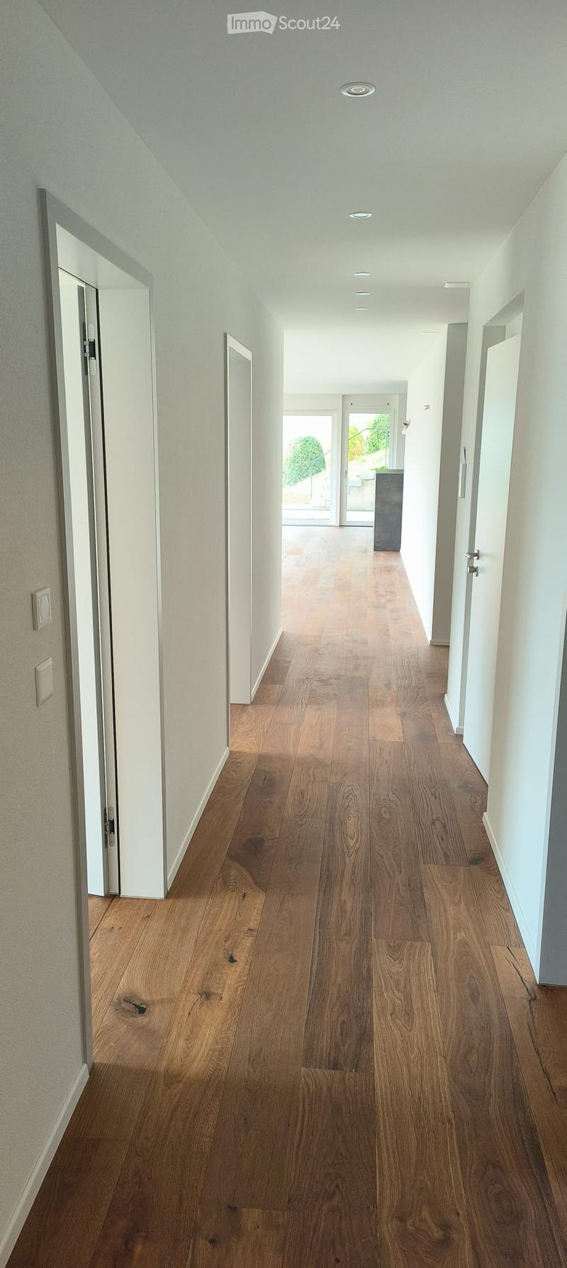 A long, well-lit hallway with wooden flooring and white walls, featuring several doors and a view to an outdoor area at the end.
