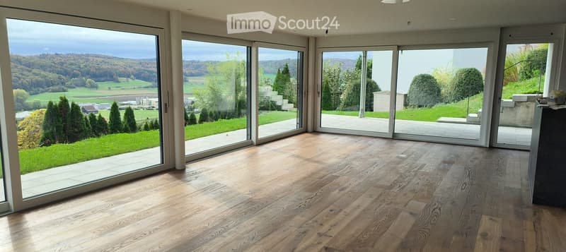 Bright, unfurnished room with large sliding glass doors opening to a landscaped garden, wooden flooring, and expansive scenic views.