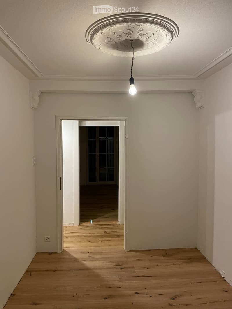 Empty hallway with wooden flooring, white walls, and a decorative ceiling light fixture. An open doorway leads to another room with glass panels.