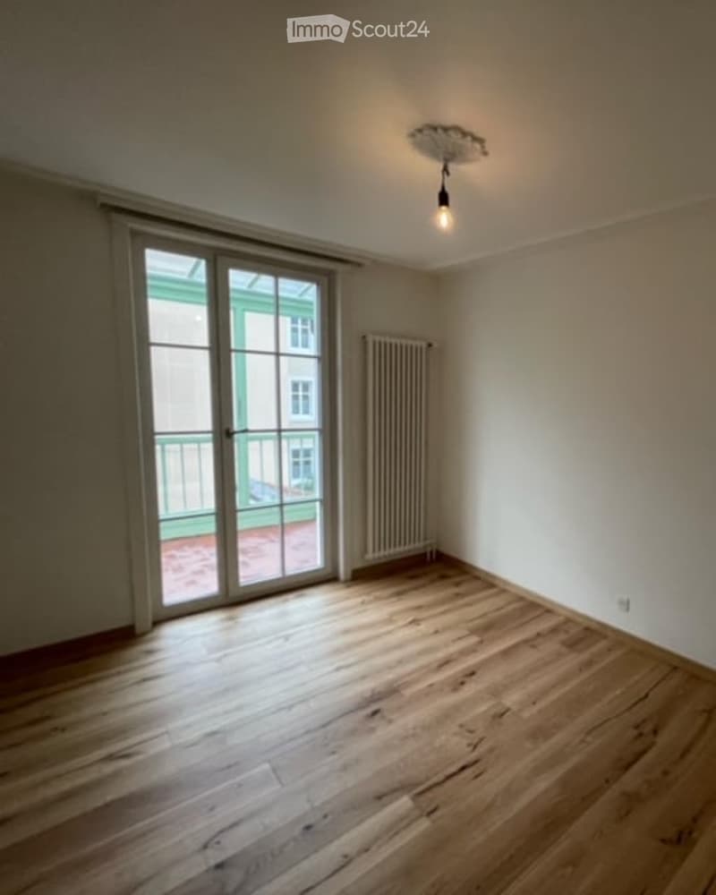 Empty room with wooden flooring, large glass sliding door leading to a balcony, radiator, and a single ceiling light fixture.