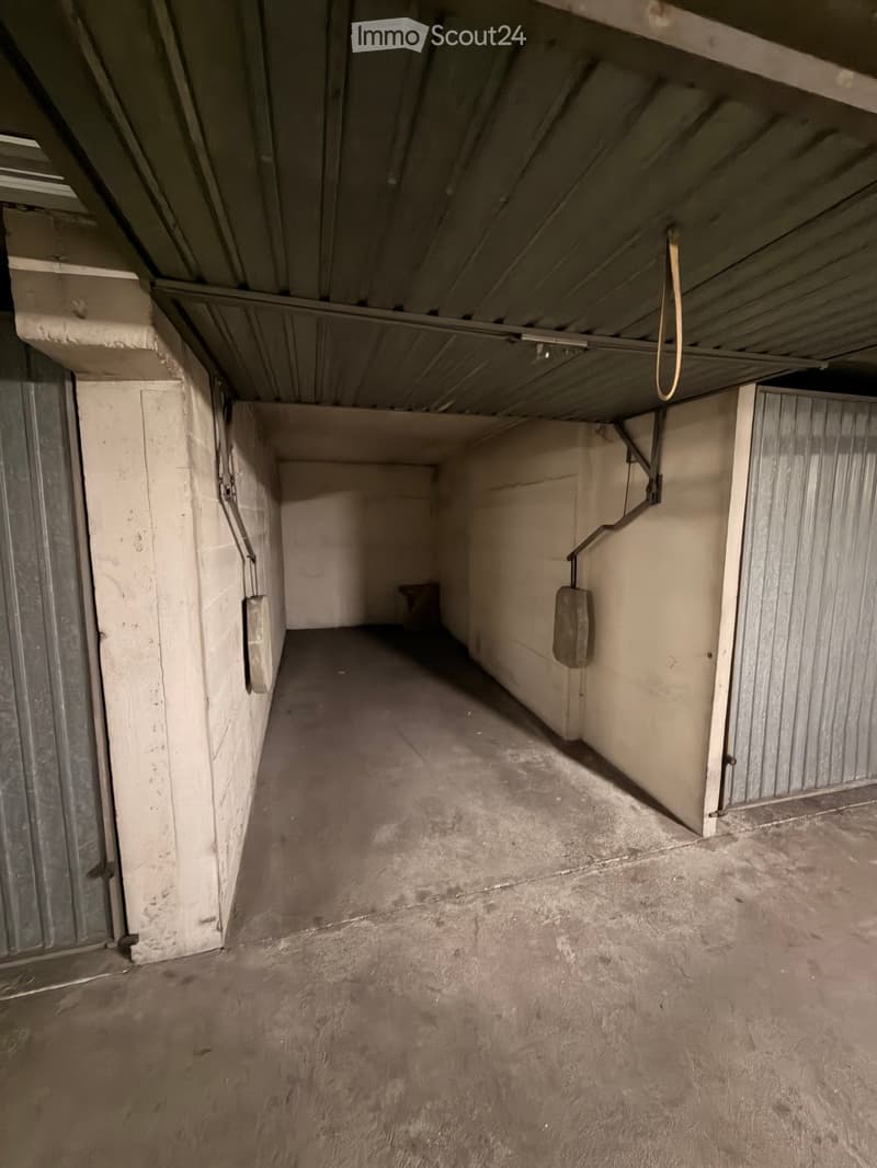 Empty garage with concrete floor, metal walls, and a metal door at the far end. Exposed pipes and a hanging cable are visible.