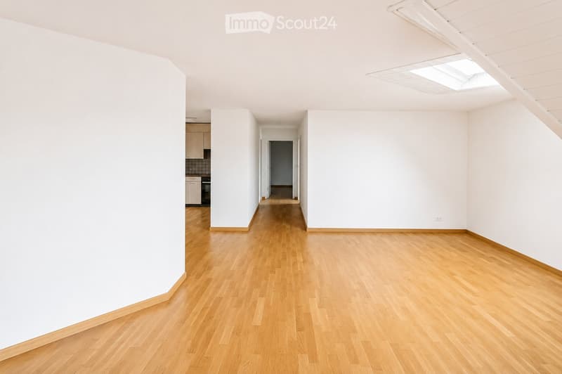 Empty room with light wooden flooring and white walls, featuring a skylight and a view of a small kitchen area in the background.