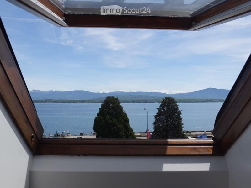 View from a balcony or terrace overlooking a lake and distant mountains, with trees in the foreground.