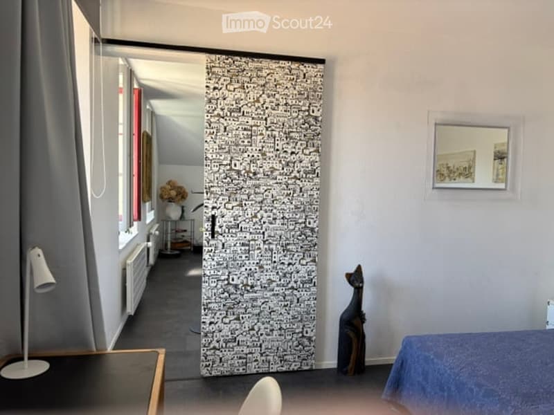 A furnished room with a bed, a lamp, a picture frame, a table, and a chair. The room has a large, patterned door and a window with curtains.