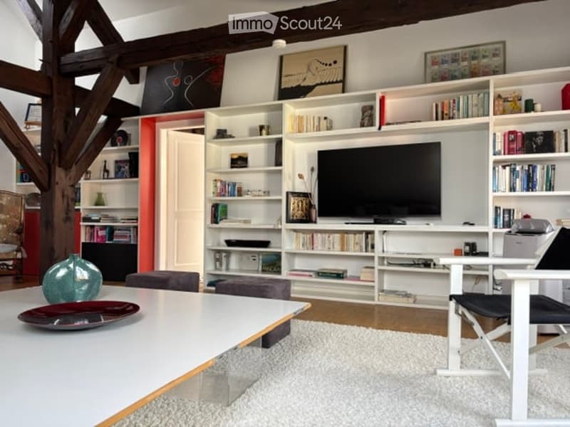 Modern living room with exposed wooden beams, white built-in bookshelves, a flat-screen TV, a white coffee table, and colorful chairs.