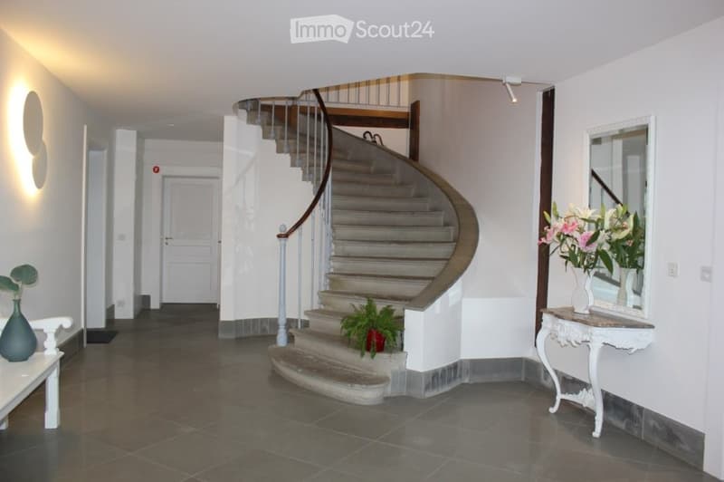 Spacious hallway with a curved staircase, white walls, dark tiled floor, and decorative vases on side tables.