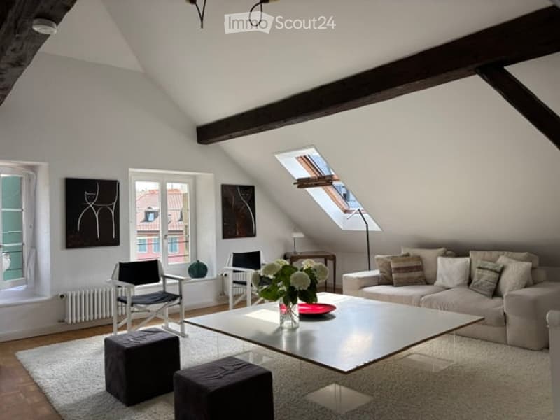 Bright living room with slanted wooden ceiling beams, large windows, and skylight. Features a grey sofa, coffee table, two black chairs, two dark grey ottomans, a small side table with a lamp, and decorative art pieces on the walls.
