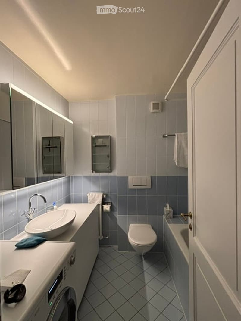 Modern bathroom with blue tiles, white sink, mirror, toilet, bathtub, and towel. Features a sleek design with integrated lighting above the mirror.