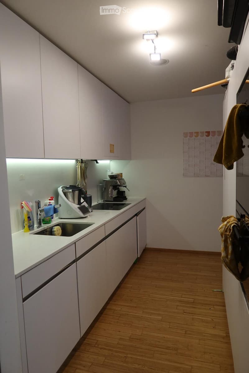 Modern kitchen with white cabinets, wooden flooring, and built-in lighting. Features a sink, coffee maker, and blender on the counter.
