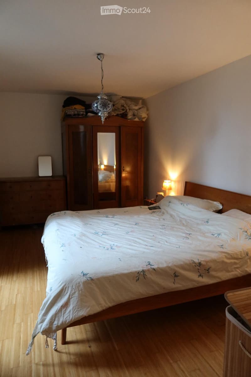 Cozy bedroom with wooden flooring, a large bed with a floral duvet, a wooden wardrobe with a mirror, a bedside lamp, and a small nightstand.