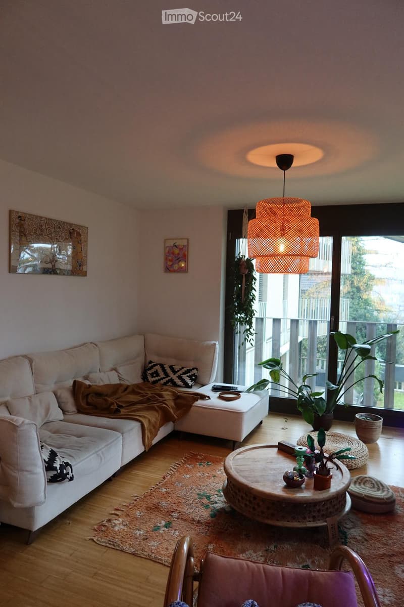 Modern living room with a white sectional sofa, a round coffee table, a pink accent chair, a patterned rug, and a large pendant light. The room features large windows with a view of a balcony and outdoor greenery.