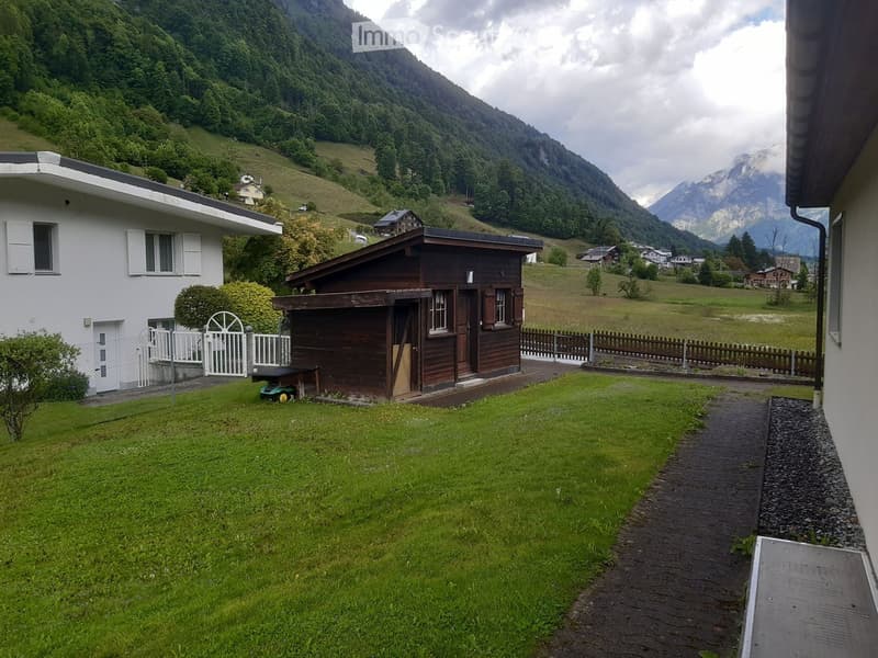 A well-maintained garden with a small wooden shed, a white fence, and a pathway leading to a house, surrounded by green hills and mountains.