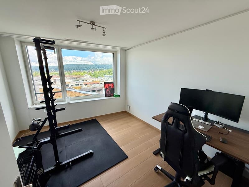 A modern, well-lit room with a large window offering a scenic view, featuring a desk with a computer setup, a gaming chair, and a fitness machine on a mat.