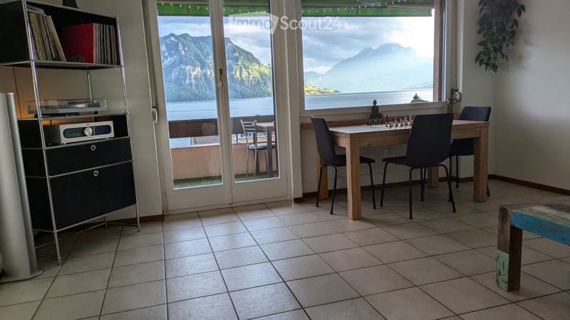 Bright room with tiled floor, large windows overlooking a scenic mountain and lake view, a wooden table with chairs, a chessboard, and a bookshelf with records and a speaker system.