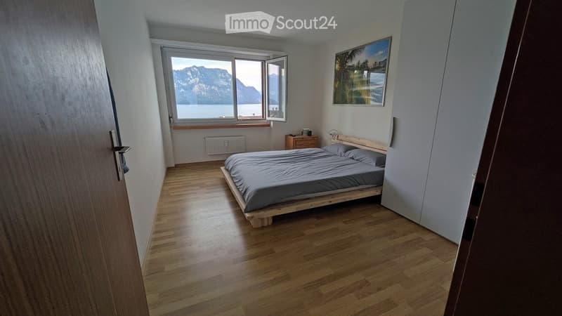 Bright bedroom with wooden flooring, a single bed, nightstand, radiator, and a scenic view of a lake and mountains through large windows.