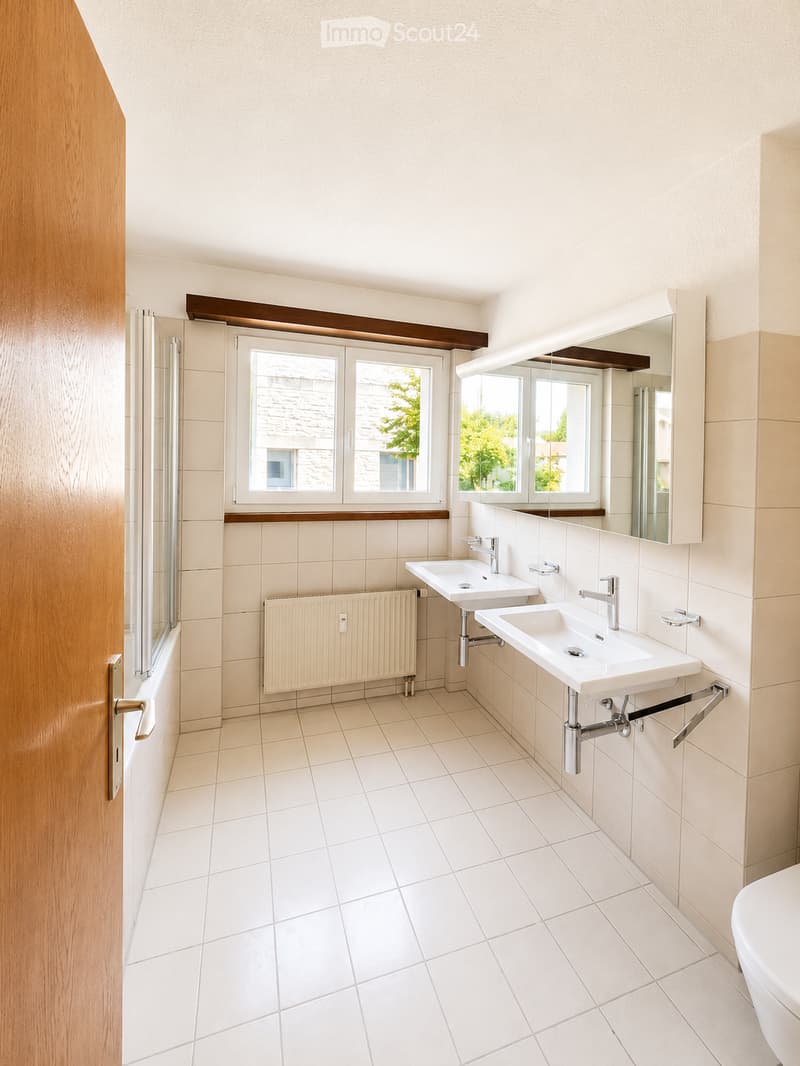 Modern bathroom with white tiled floors and walls, two sinks with modern faucets, a large mirror, a radiator, and a partially visible toilet.