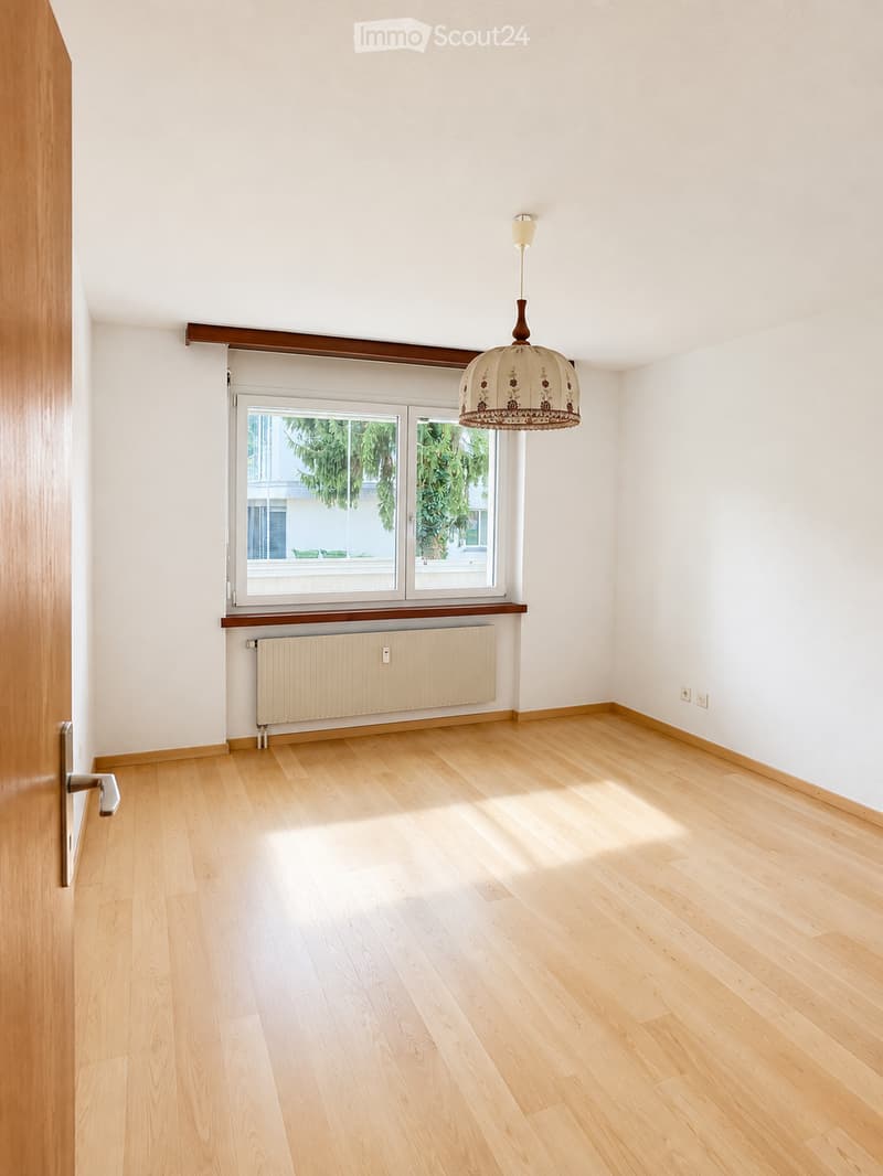 Empty room with light wooden flooring, white walls, a window with a view of greenery, a radiator, and a decorative hanging light fixture.