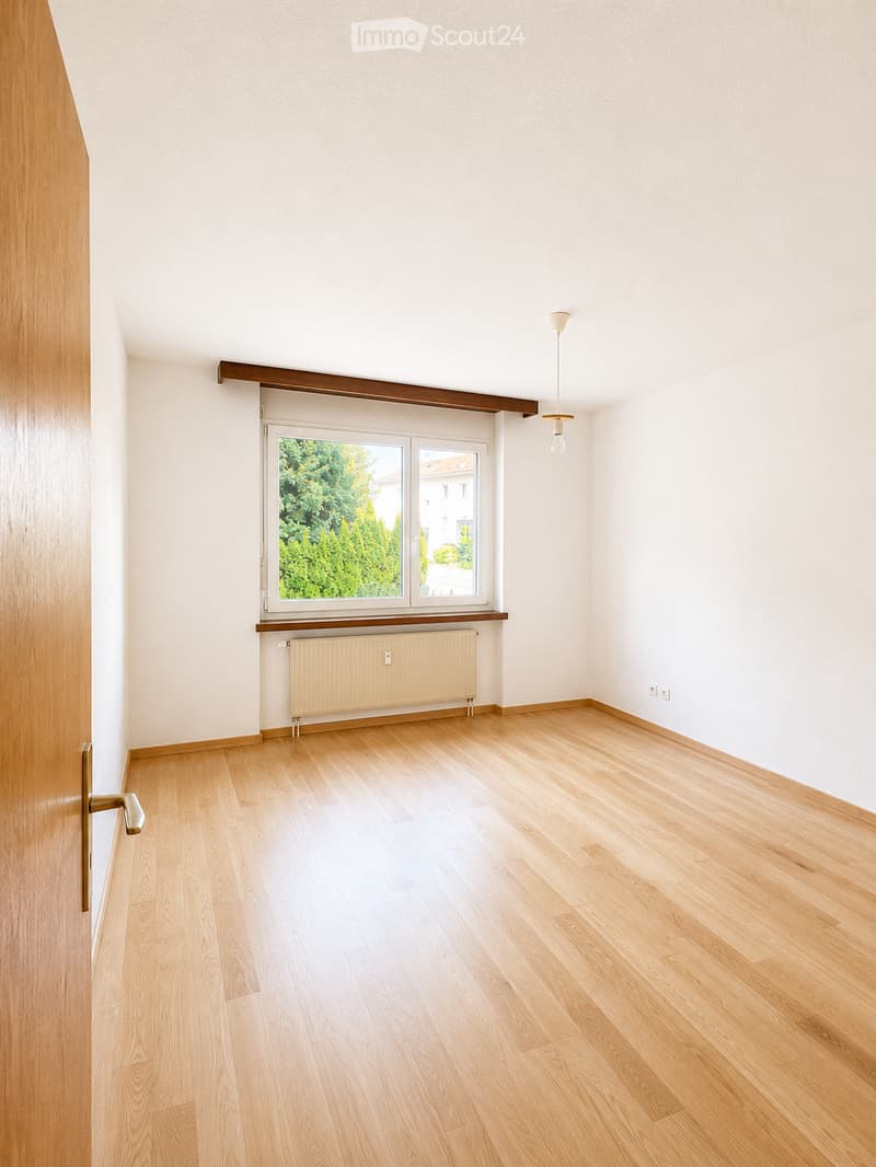 Empty room with light wooden flooring, white walls, a window with a view of greenery, a radiator, and a hanging light fixture.