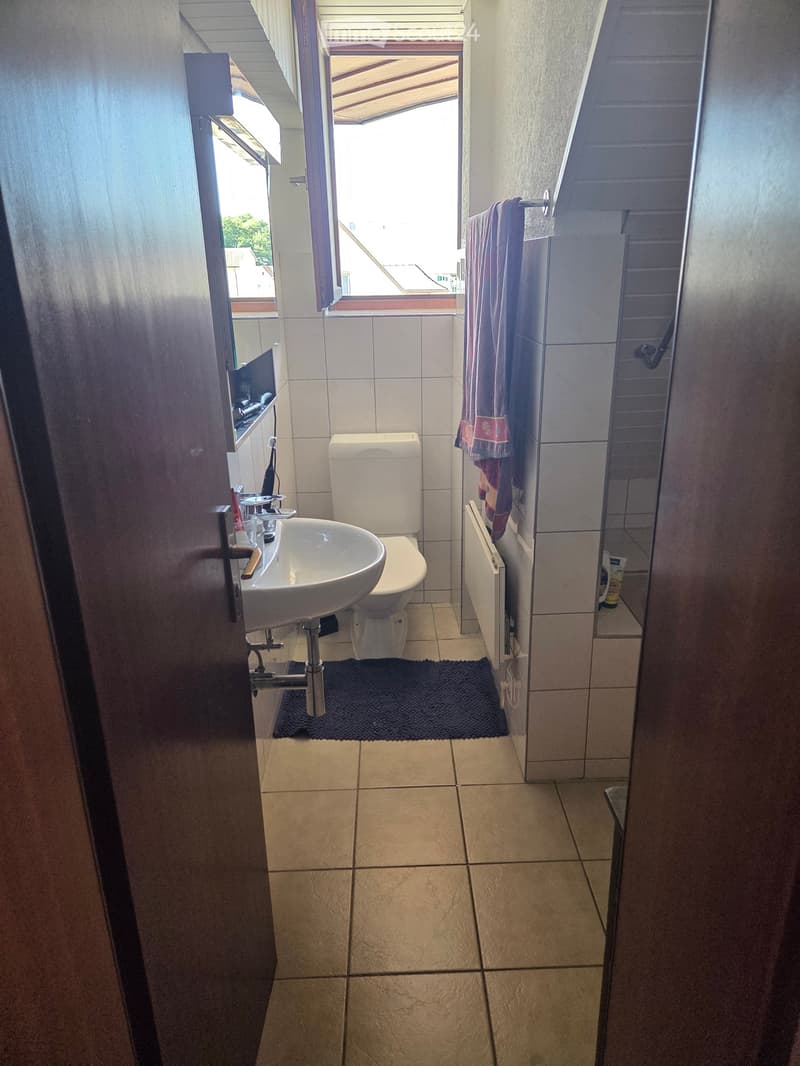 Bathroom with a sink, toilet, shower, and a towel hanging on a rack. The floor is tiled, and there is a small window with blinds above the shower area.