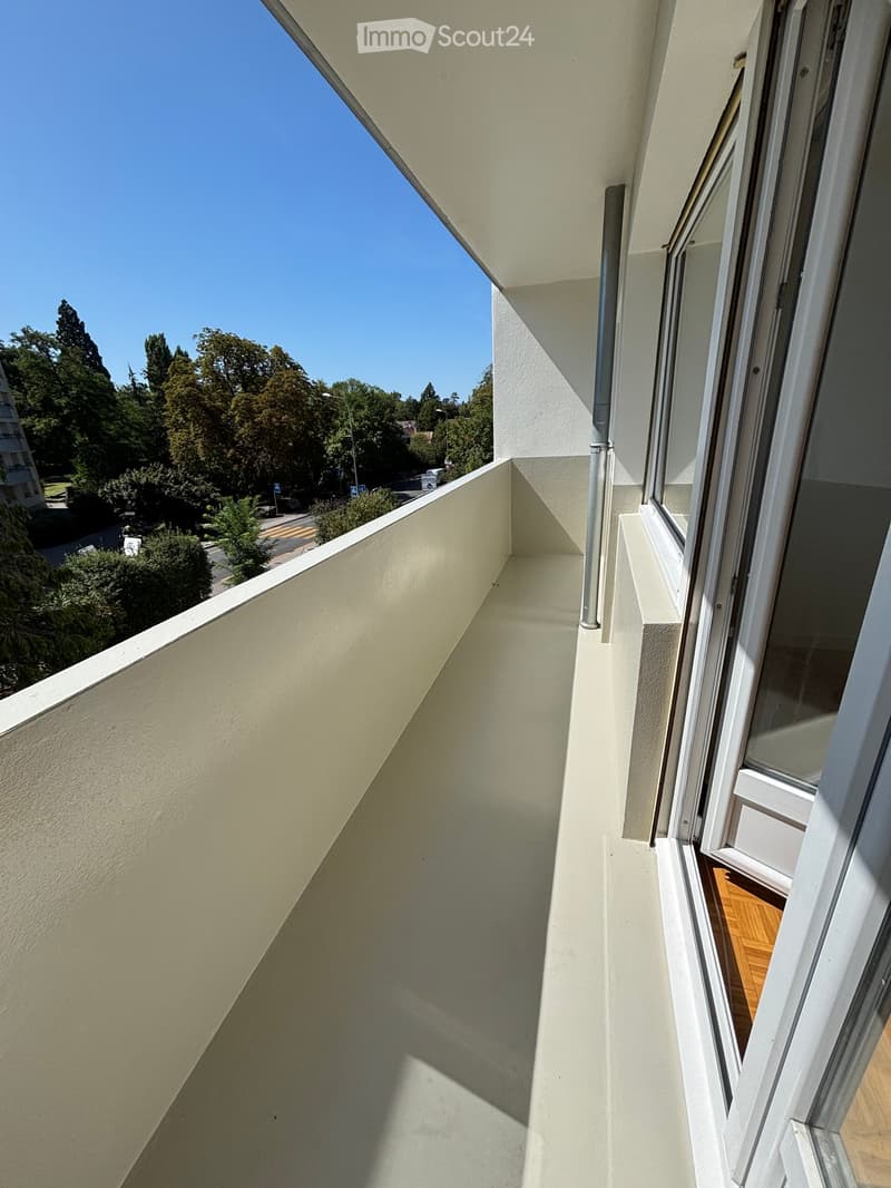 Balcony with white walls, large windows, and a view of trees and a street.