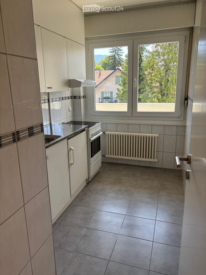 Modern kitchen with white cabinets, tiled floor, oven, radiator, and large windows overlooking a green area.