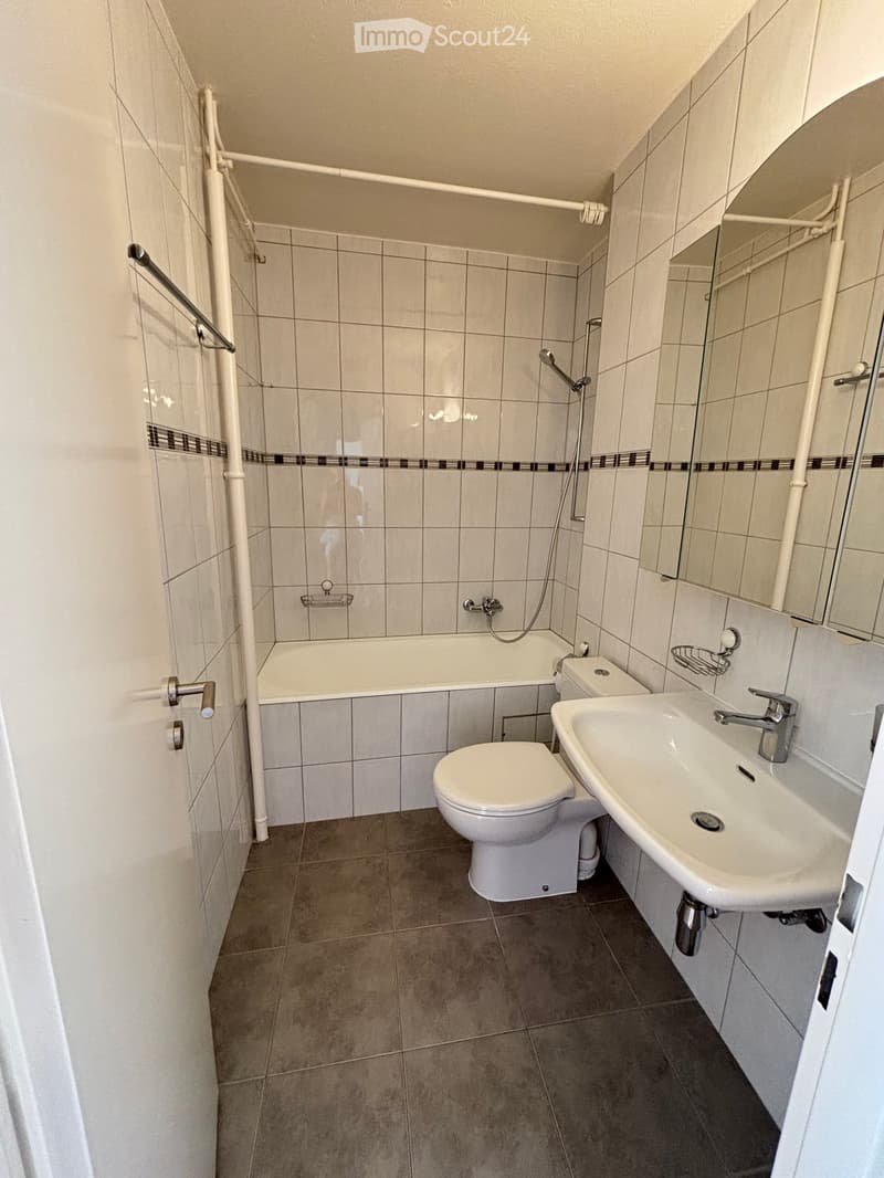 Modern bathroom with a bathtub, toilet, sink, and mirror. Features tiled walls and a grey tiled floor.