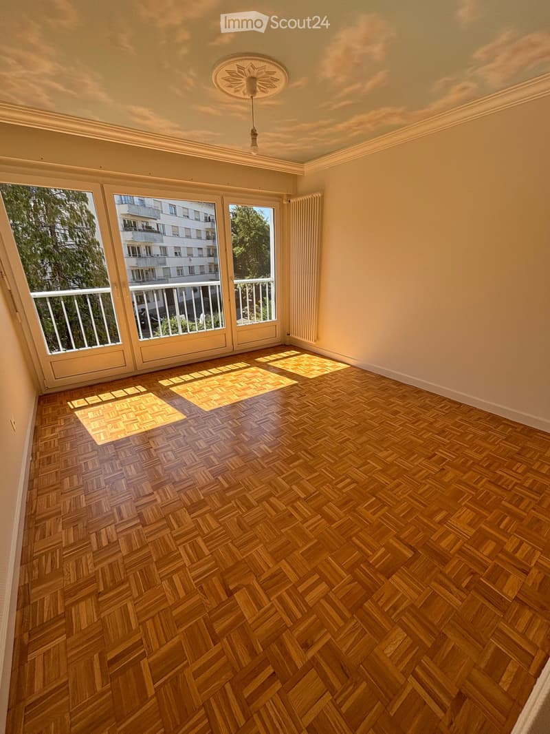 Empty room with wooden herringbone flooring, large windows with white frames, and a radiator on the wall.