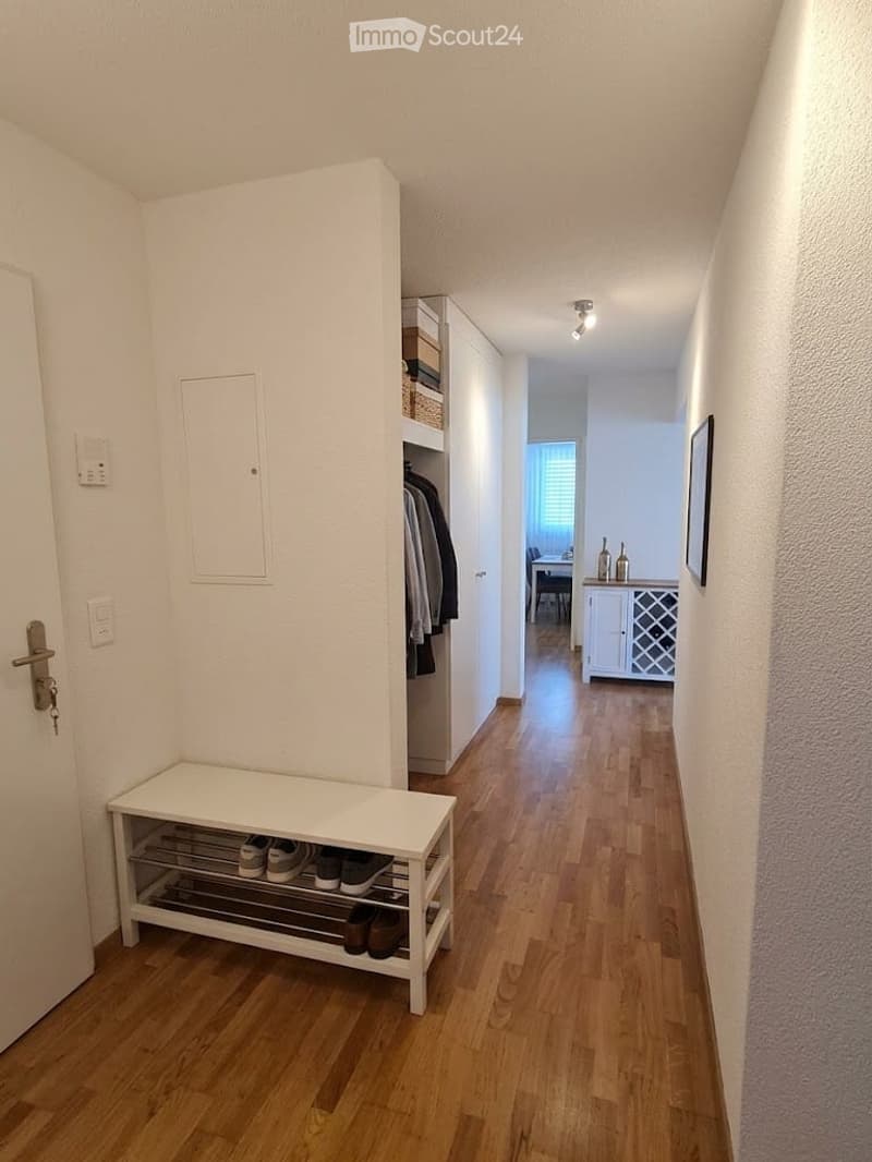 A well-lit hallway with wooden flooring, white walls, and a built-in shoe rack. A small bench with shoes is visible, and a closet with hanging clothes is on the right side. The hallway leads to another room with a dining area visible in the background.