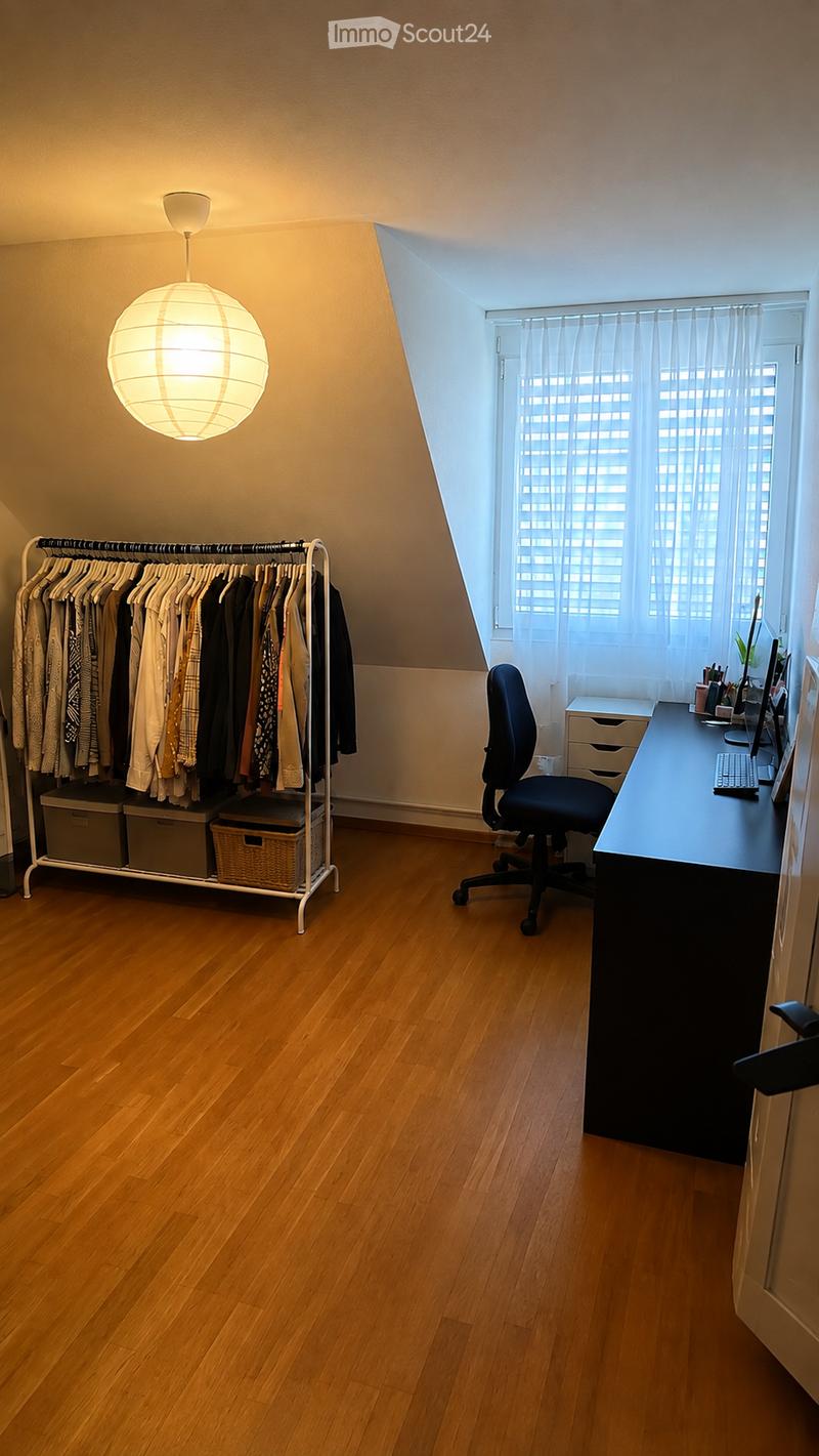 A well-lit room with wooden flooring, a large window with blinds, a desk with a chair, a clothing rack with hanging clothes, and storage boxes.
