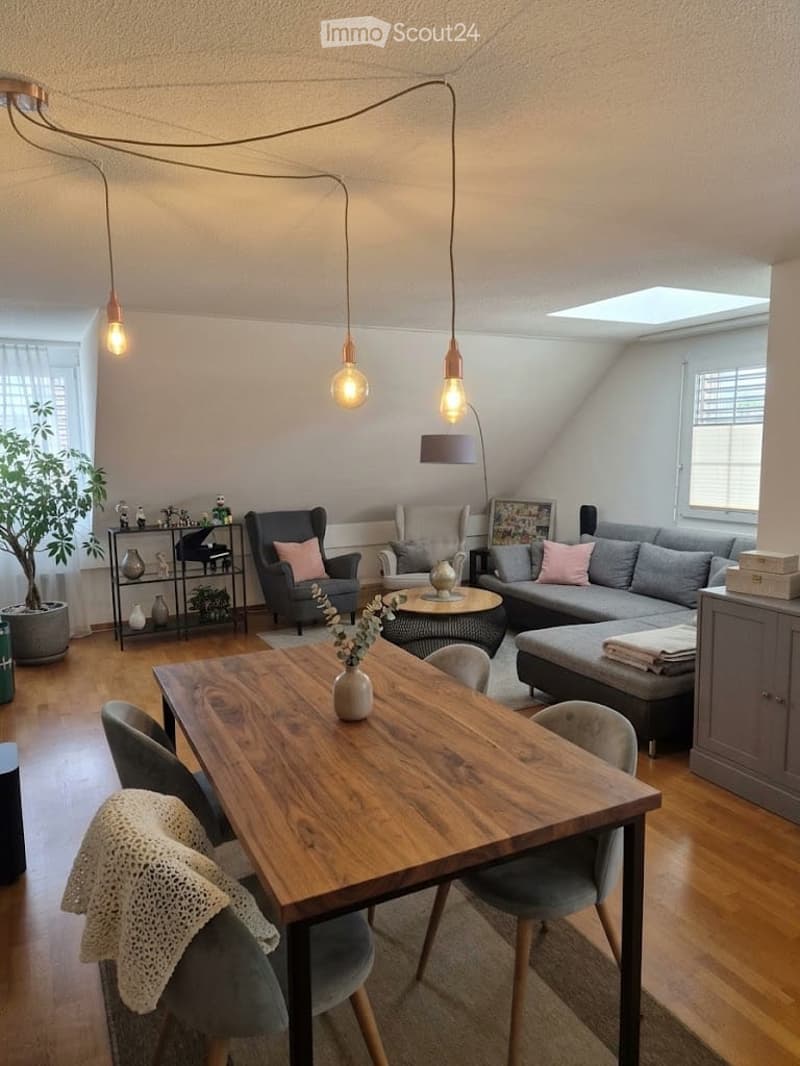 Bright, open living and dining area with wooden flooring, modern lighting, grey sofa, armchair, coffee table, dining table with chairs, and decorative shelf with vases and plants.