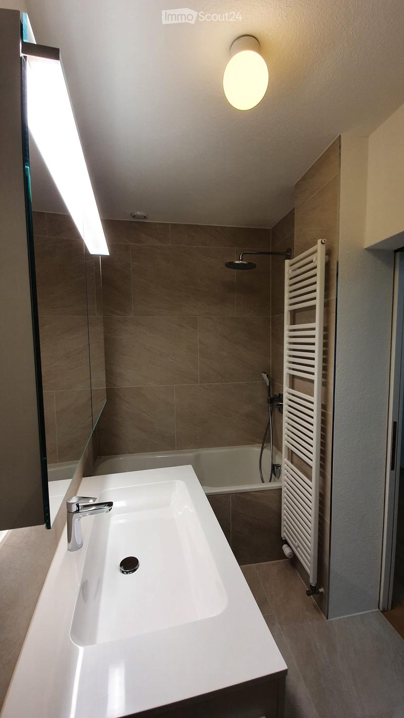 Modern bathroom with a white sink, chrome faucet, bathtub, showerhead, and a white radiator. The walls are tiled in beige, and the ceiling has a round light fixture.