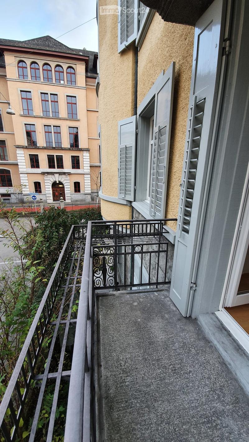 Balcony with grey concrete floor, black metal railing, open shutters, and greenery visible below.