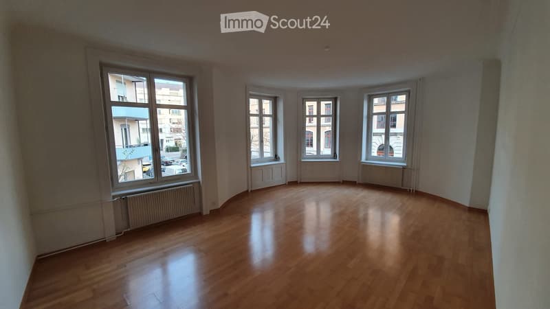 Empty room with wooden flooring and large windows, featuring radiators under the windows.