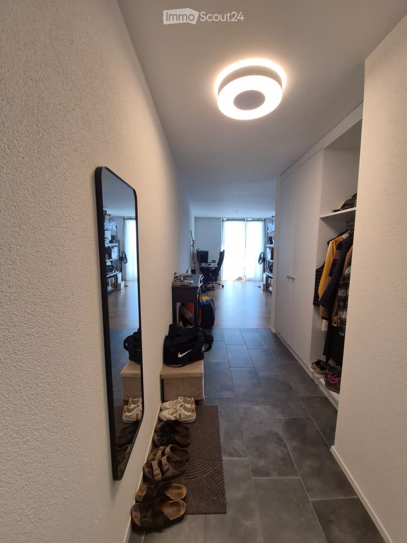 A narrow hallway with a round ceiling light, a built-in closet with hanging clothes, a mirror, and a small bench with bags and shoes.