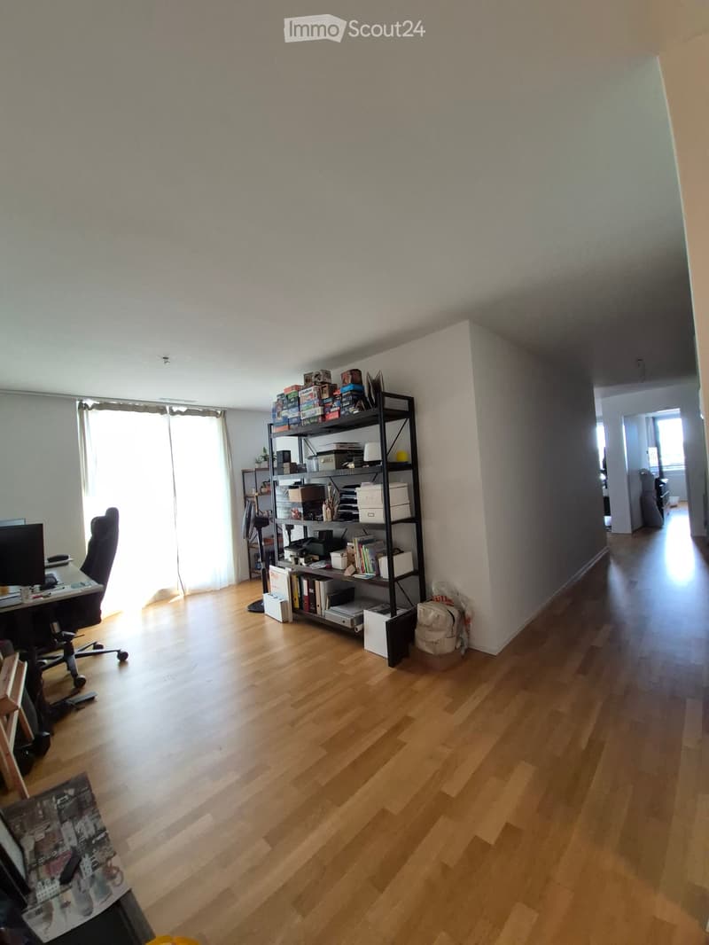 A well-lit room with wooden flooring, a black metal shelving unit filled with books and boxes, a desk with a chair, and a large window with white curtains.