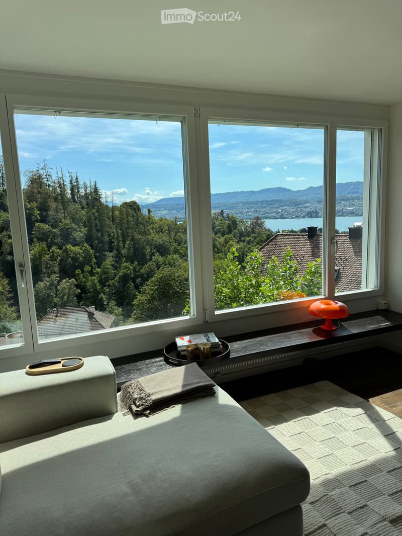 Bright living room with large windows offering scenic views, featuring a grey sofa, a small table with a lamp, and decorative items.