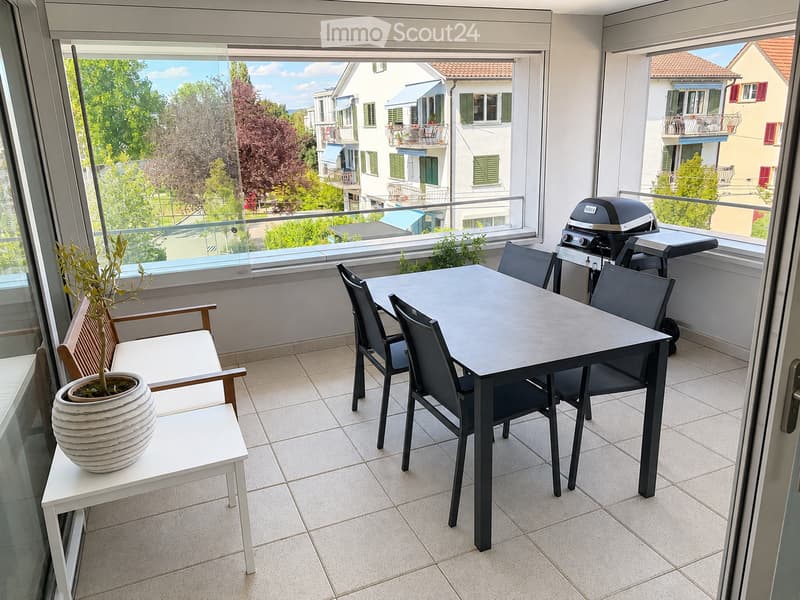 Balcony with a dining table, chairs, a bench, a potted plant, and a grill, overlooking a scenic view of neighboring buildings and greenery.