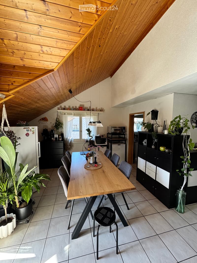 A cozy, well-lit dining area with a wooden table and chairs, surrounded by modern cabinets, a refrigerator, and various kitchen appliances. The space features a slanted wooden ceiling and large windows allowing natural light to fill the room.